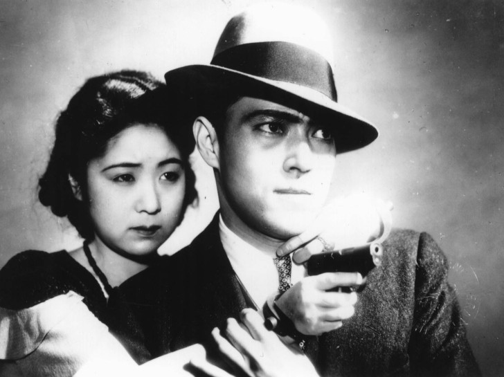 close-up of a woman behind a man with a fedora, she has one hand on his shoulder and the other points a gunalr