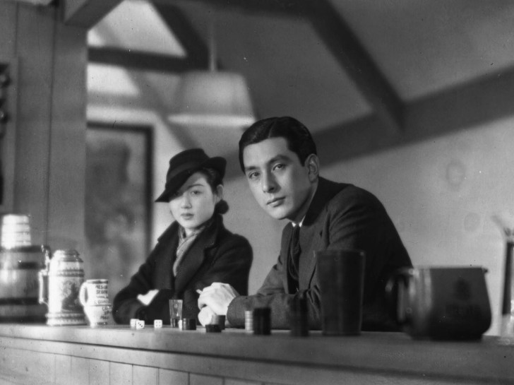 a young Japanese man and woman sit next to one another at a bar looking at the vieweralr