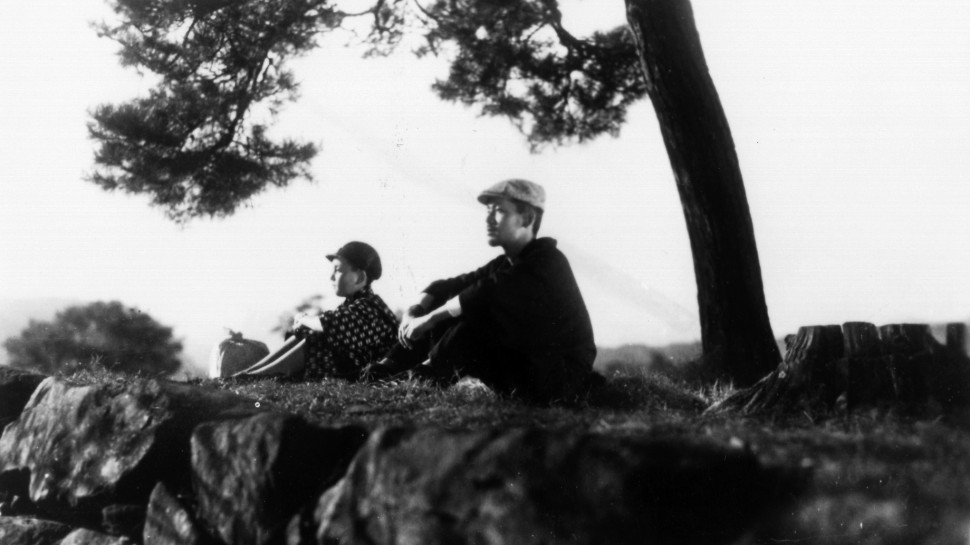 wide shot of a young boy and older man sitting serenely next to a cliff and a lone treealr