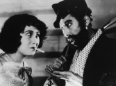 a woman with a cigarette looks provocatively at a bearded man with a pipe