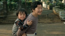 an older Japanese woman outside carrying a young child on her back, both are smiling