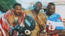 two bloody Black men, including one holding a film camera, help a third to walk