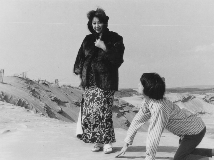 a woman smiling and clutching her coat as she looks down at a kneeling man on the desertalr