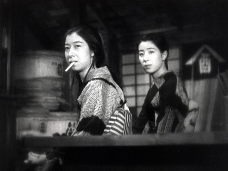 two women in traditional Japanese dress stand next to each other, bodies facing sideways and turned toward the camera, one with a cigarette hanging out of her mouthalr