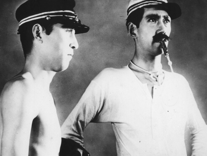 a young man without a shirt and a military hat on stares blankly next to a mustachioed man with a whistle in his mouthalr