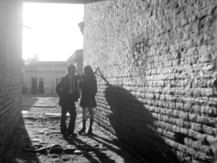 a wide shot down an alley of a man in a suit talking to a woman in a dress and boots  while their shadows stretch across the brick wallalr