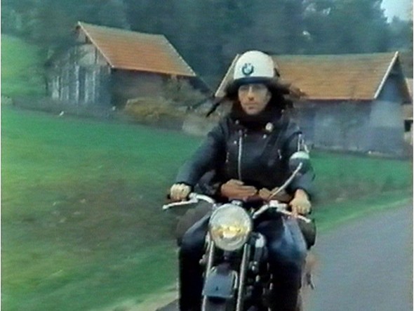 white man with long hair and a helmet rides a motorcycle through the countrysidealr