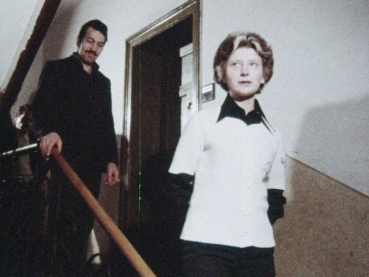 a white woman walks down the stairs inside an apartment building, looking at the camera, with a white man close behind heralr
