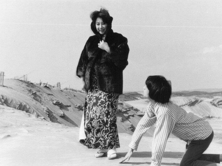 a Japanese woman standing on the beach, in a fur coat, smiling and a Japanese man on his hands and knees before heralr