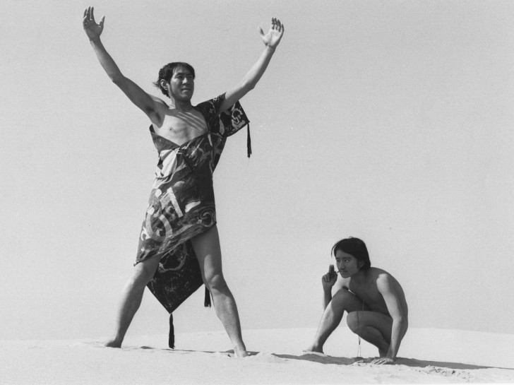 two Japanese men in the desert: one, wearing a tribal-looking garment with one shoulder bare, raises his arms high; the other, nearly naked, crouches next to himalr