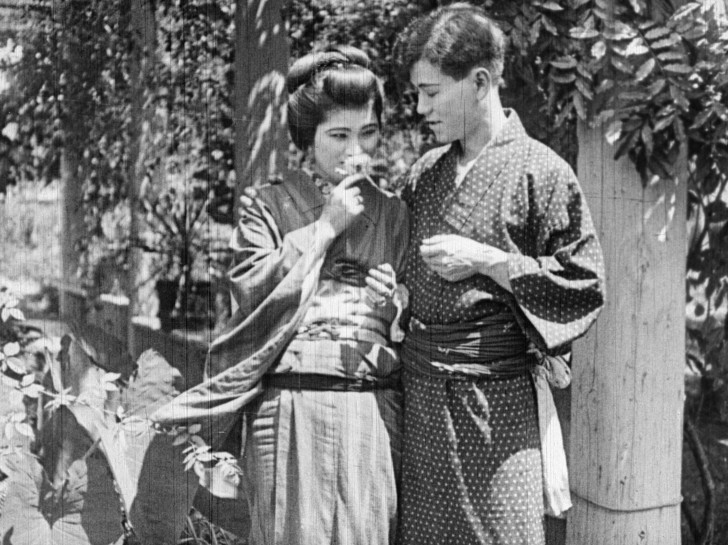 a young Japanese man and woman stand close to one another while she smells a floweralr