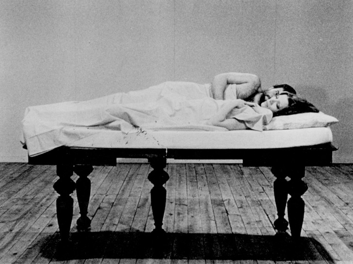 a white man and woman, covered by a sheet, lying on a mattress on a table in an empty roomalr