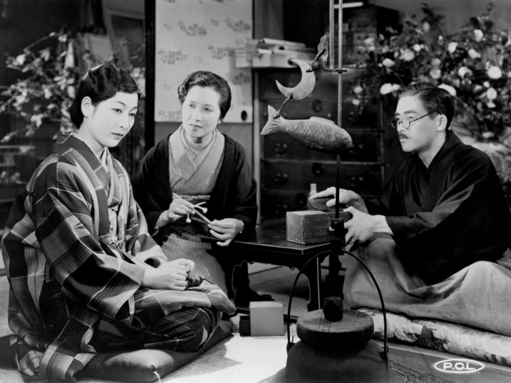 an older Japanese man and a woman sit around a table and fish sculpture staring eagerly at a younger woman who looks awayalr