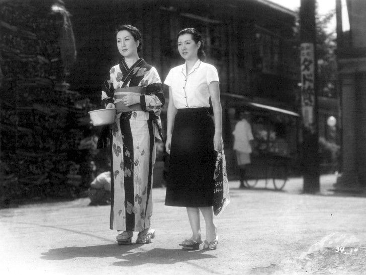 Miura Mitsuko in traditional Japanese dress, carrying a bowl, walks alongside Takamine Hideko in a western skirt and blousealr