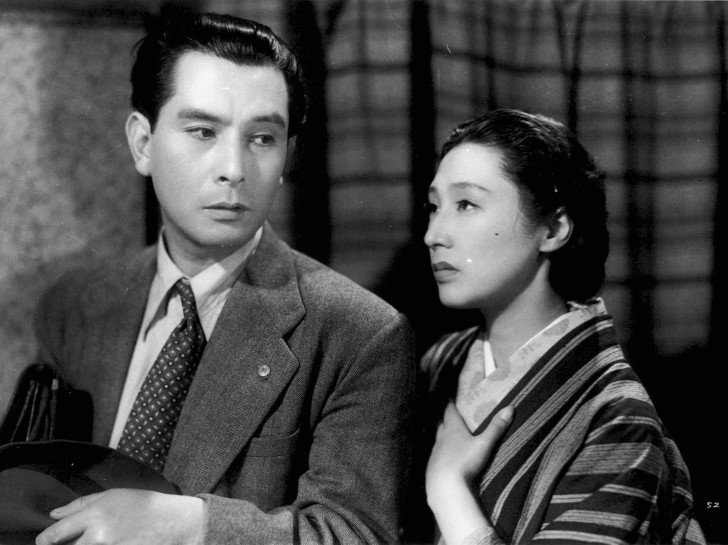 a despondent Takamine Mieko stands next to a distracted Uehara Ken, with her hand on her heart alr