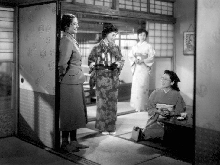 a standing Hara Setsuko laughs with a seated woman while another woman carries bottles of winealr