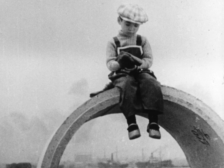 a small boy in a plaid cap, sits on a large arched structure outsidealr