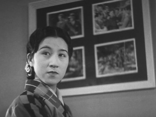 standing in front of pictures on the wall, a defiant looking Kurishima Sumiko gazes to the side with pursed lipsalr