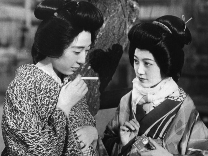 one young Japanese women holds out her cigarette to the other who has matchesalr