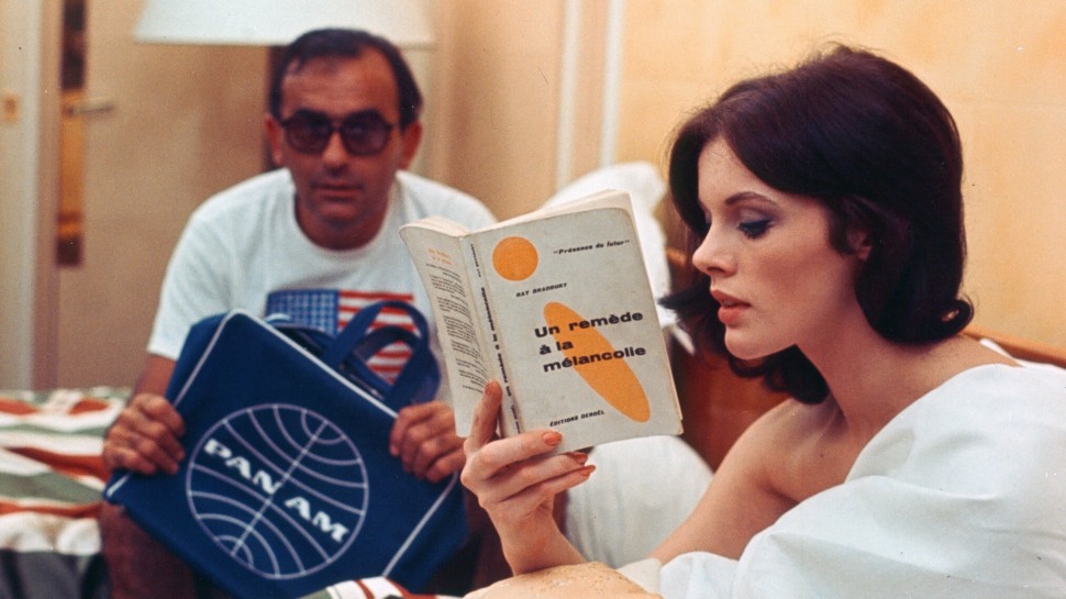 a woman sitting up in bed reading a book about sadness while a man sits across from her with a PanAm travel bagalr