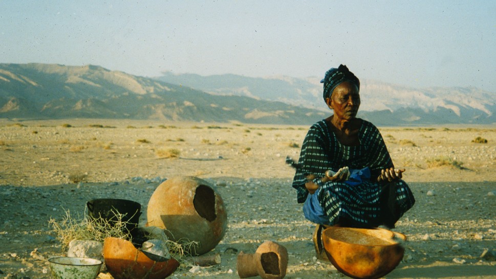 An older woman sitting near pottery vessels in the desert with her eyes closed and hands on her lap, facing upalr