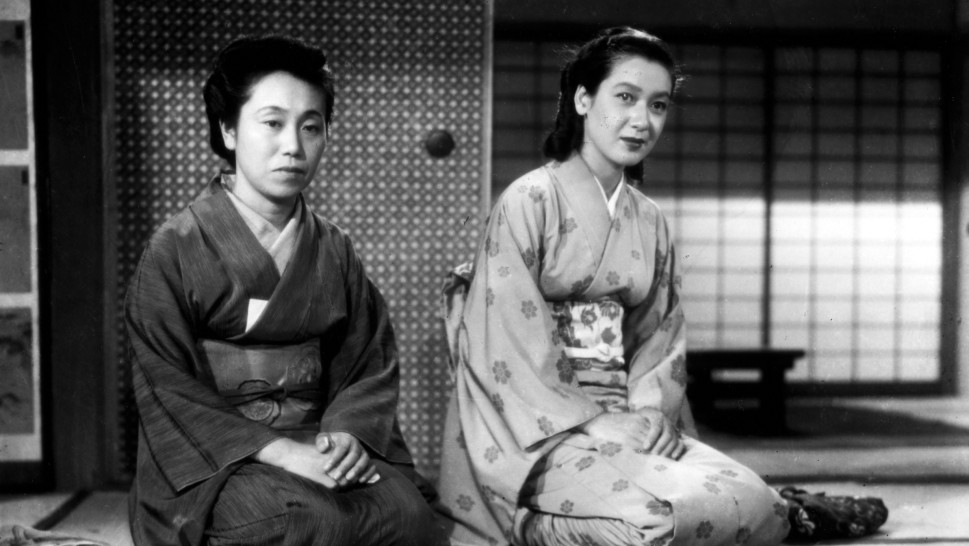 two women in traditional Japanese dress sitting on a mat indoors, looking at the cameraalr