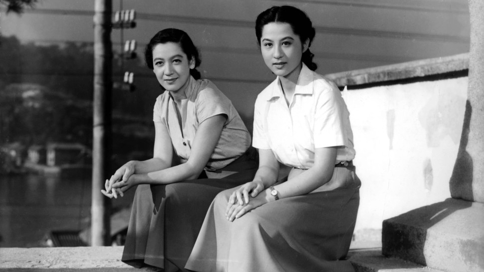 Two young Japanese women in shirts and skirts sit on stools on the street, slightly smiling and looking at the vieweralr