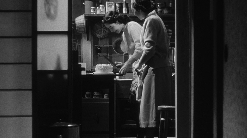 Medium wide shot, down a short hallway, of two women in the kitchen, one cutting a cake  and smilingalr