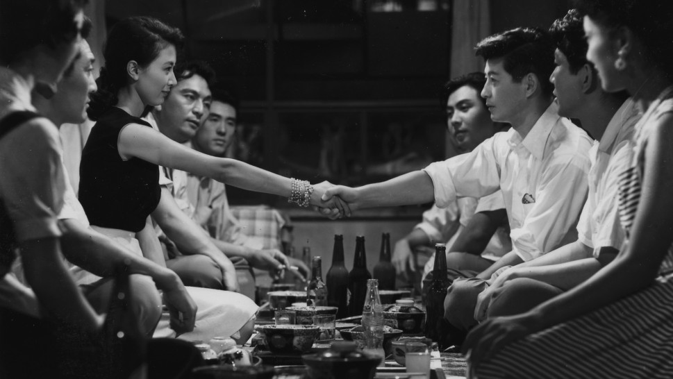 a group of young Japanese men and women at dinner gaze at one man and a woman shaking hands over the tablealr