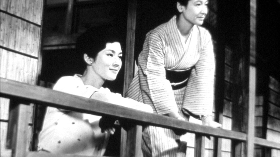 a young woman in Western-style dress and an older woman in traditional Japanese dress lean on a porch railing, smiling and looking out alr