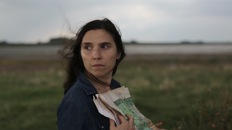 a young Argentinian woman in a field, holding a map and looking awayalr