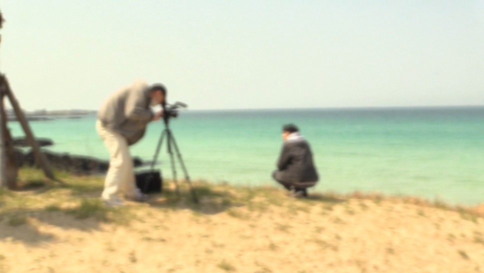 out-of-focus scene of a man filming on a beach while another sits in the sandalr