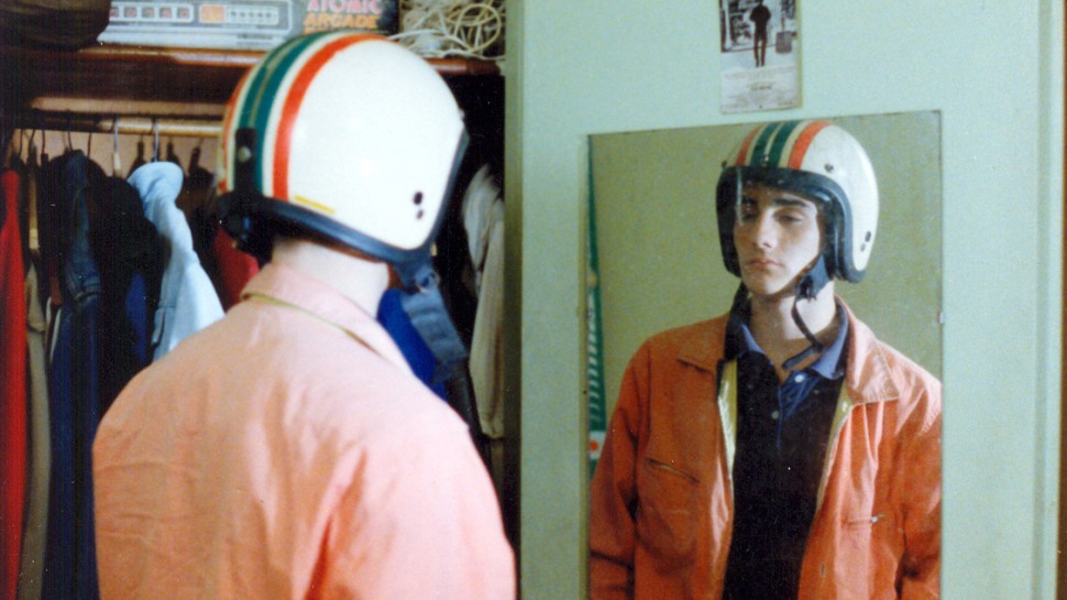 a young white man wearing a striped motorcycle helmet looks in the mirroralr