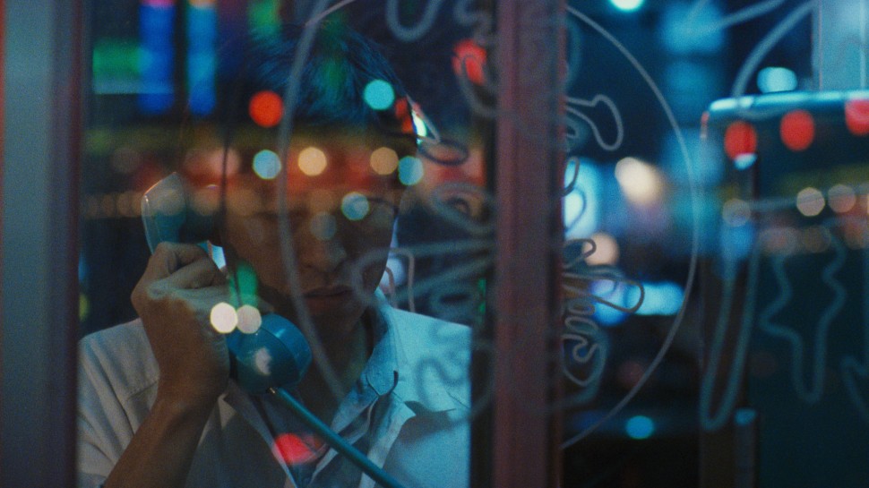 a young Asian man talks in a phone booth with colorful reflections of lights on the glassalr