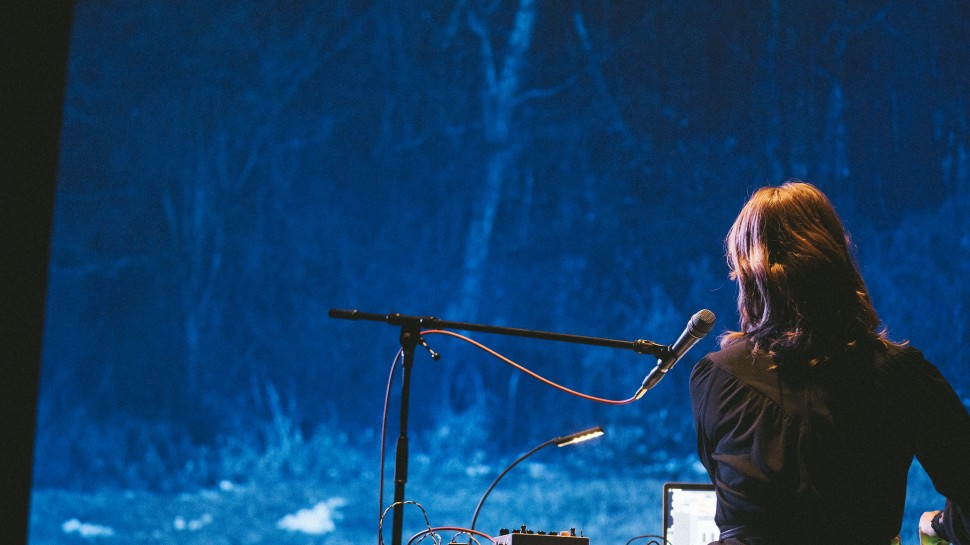 the back of Ishibashi Eiko with a microphone and electronics in front of a screen featuring a forested landscapealr