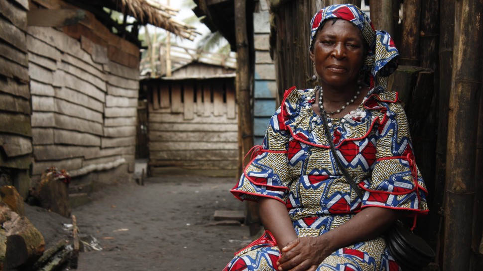 an older African woman in a brightly patterned dress sits outside, looking directly at cameraalr