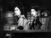 two women in traditional Japanese dress stand next to each other, bodies facing sideways and turned toward the camera, one with a cigarette hanging out of her mouth