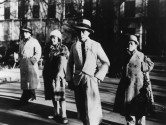 three men and two women in 30s-era wear stand in the street with serious expressions