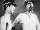 a young man without a shirt and a military hat on stares blankly next to a mustachioed man with a whistle in his mouth