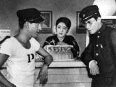 a young man with a military cap leans on the counter of a bar looking at another man in military garb looking down while a woman stands behind the bar, between them