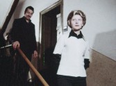 a white woman walks down the stairs inside an apartment building, looking at the camera, with a white man close behind her