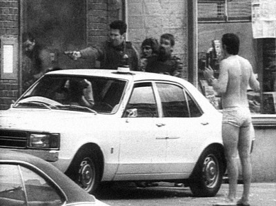 a white man in his underwear stands on one side of a car in the street holding his hands up while a white man on the other side shoots a gun