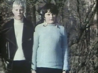 two young white boys stand next to one another in front of a forest of trees