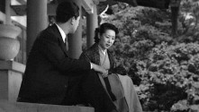 man in a suit sitting on the stairs of a Japanese temple with his head turned toward the woman next to him in traditional Japanese dress, speaking