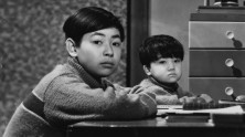 two young Japanese boys sit sideways at a desk in a bedroom turned toward and looking at the viewer