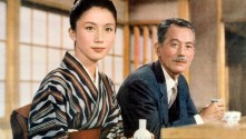 a young Japanese woman in traditional dress and a Japanese man in a suit sit at a bar, faintly smiling, looking out at the viewer