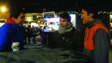 three young Afghan men sit around a table next to a food stall, smiling and talking, on a cold winter night