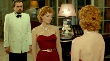 Delphine Seyrig ins a red, strapless dress looking in the mirror, with a male costar in a tuxedo behind her, both serious