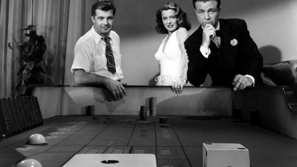 Director Robert Rossen with Ellen Drew and Dick Powell at the end of a craps table looking at oversized dicealr