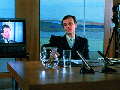 actor at desk with microphones in front and tv monitor in back